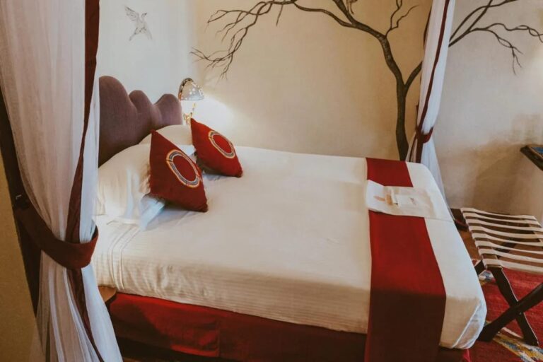 Un grand lit aux coussins rouges entre deux rideaux blancs, devant un arbre peint au mur