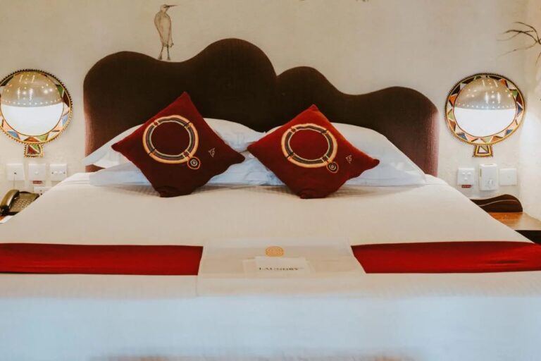 Deux coussins rouges brodés sur un grand lit, sous un héron peint au mur et entre deux appliques colorées