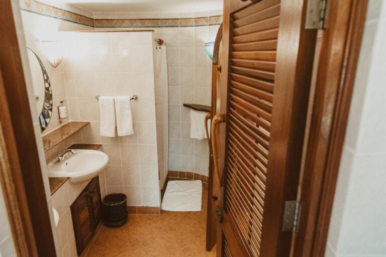 Une salle de bains vue depuis sa porte en bois à persiennes, avec un lavabo et une douche à rideau