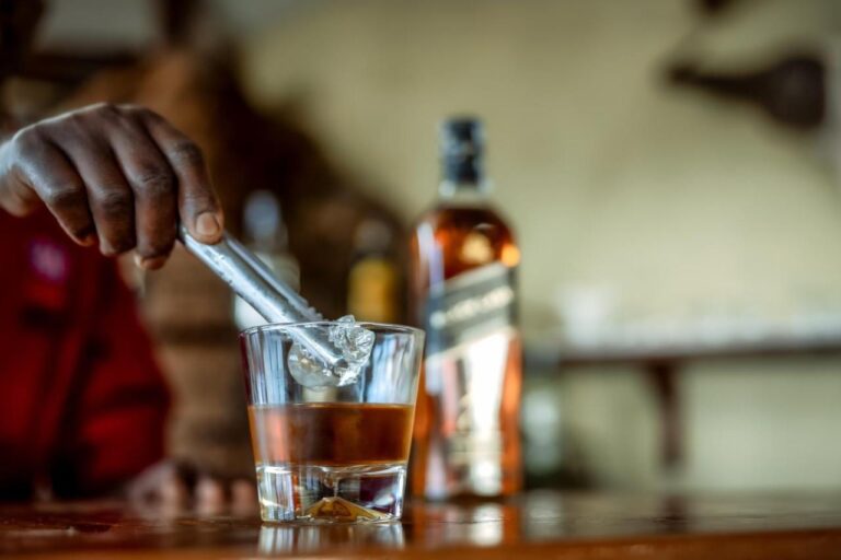 Une main dépose un glaçon dans un verre de whisky, sur le comptoir du bar