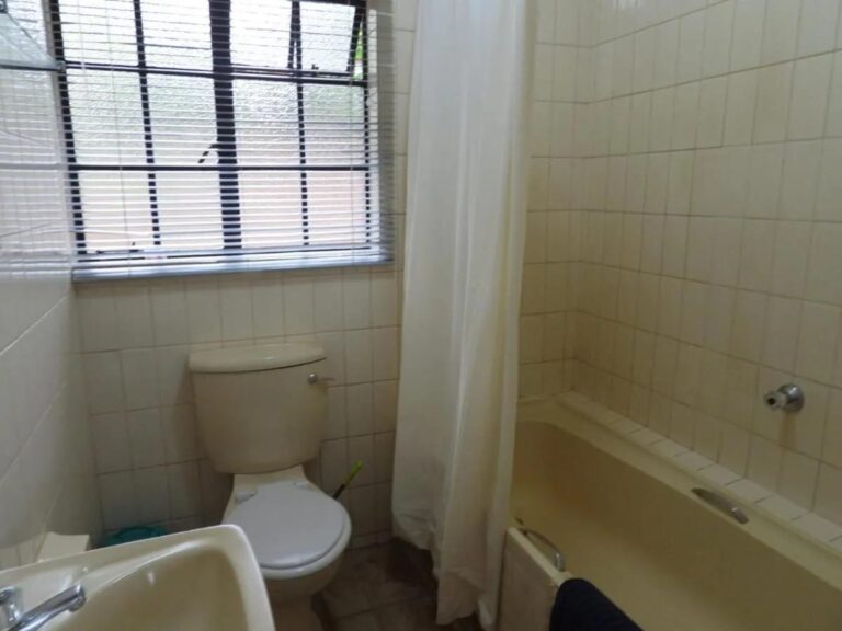 Une salle de bains carrelée, avec baignoire à rideau, toilettes et lavabo, sous une fenêtre à store