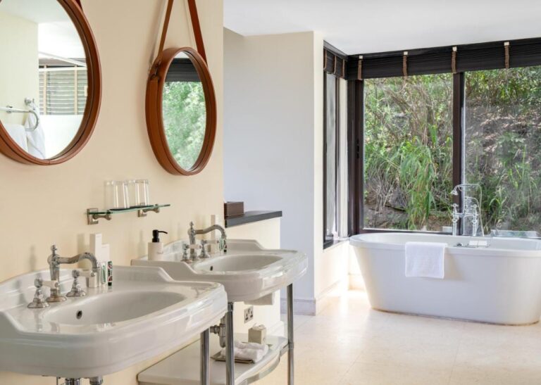 Une salle de bains avec deux lavabos sur pied, des miroirs ronds et une baignoire devant une baie vitrée