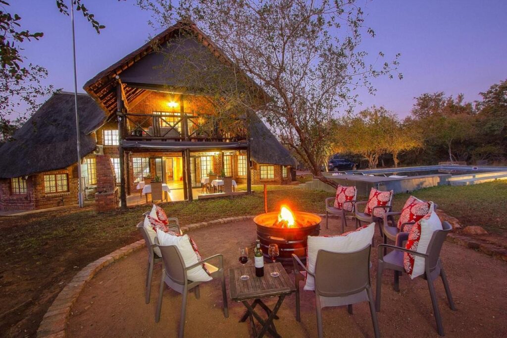 Le bâtiment en brique et chaume de l'Imbube Safari Lodge au crépuscule, avec un brasero allumé entouré de fauteuils, près de la piscine