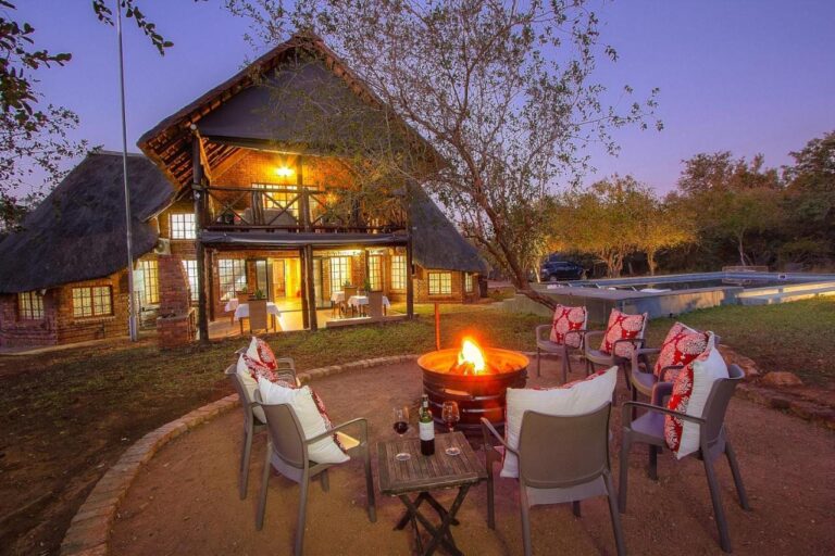 Le bâtiment en brique et chaume de l'Imbube Safari Lodge au crépuscule, avec un brasero allumé entouré de fauteuils, près de la piscine