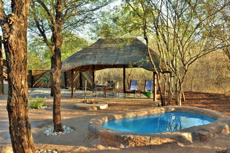 Une piscine ronde devant un abri au toit de chaume, avec une table à manger et deux transats rayés