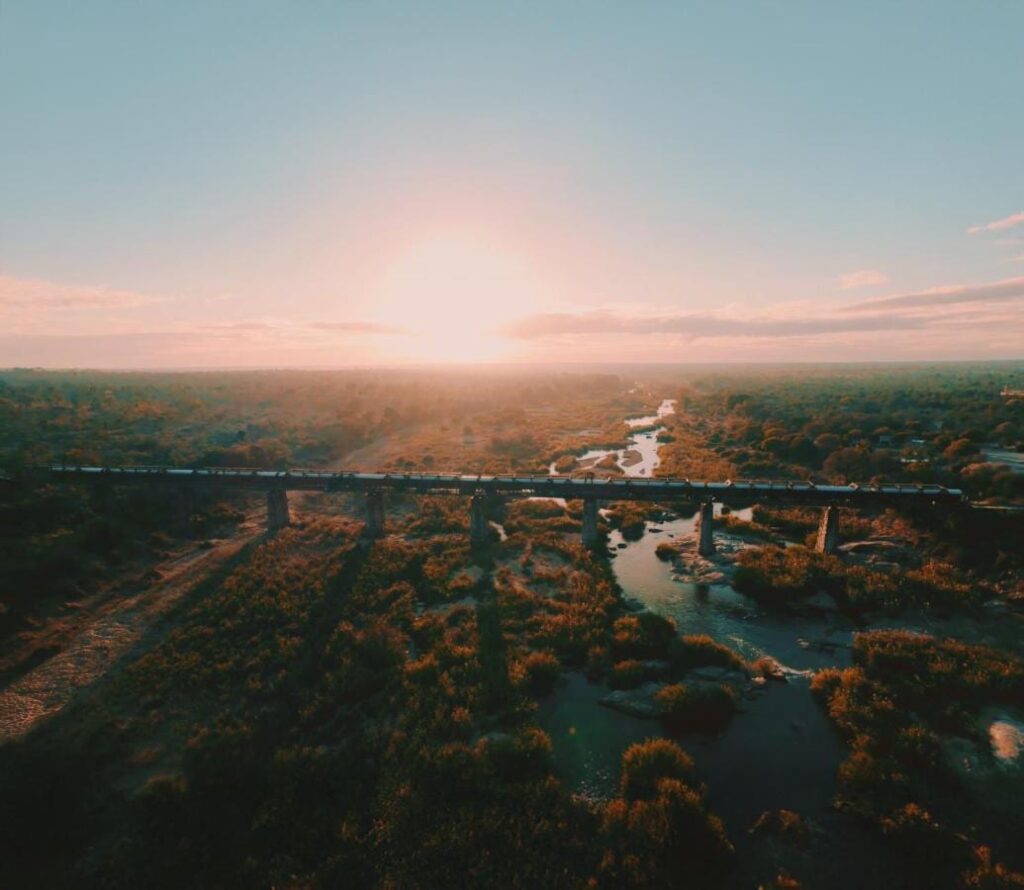 Le train du Kruger Shalati sur le pont de la Selati, vu du ciel, au-dessus de la rivière et de la brousse, le soleil bas sur l'horizon