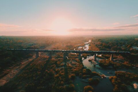 Le train du Kruger Shalati sur le pont de la Selati, vu du ciel, au-dessus de la rivière et de la brousse, le soleil bas sur l'horizon
