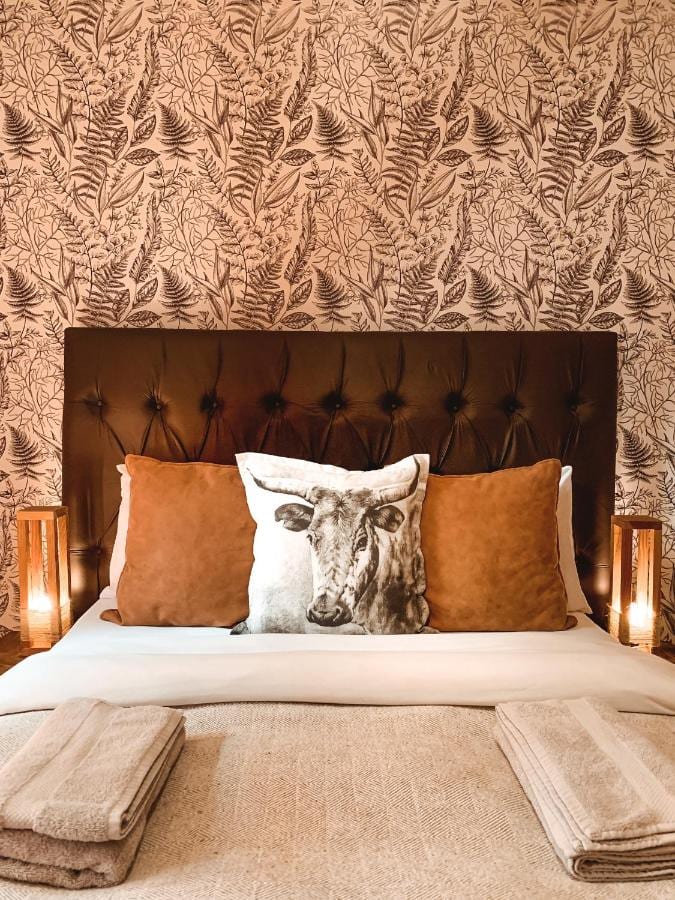 Un grand lit à tête de lit capitonnée, avec des coussins cognac et un coussin à tête de vache, devant un papier peint à fougères