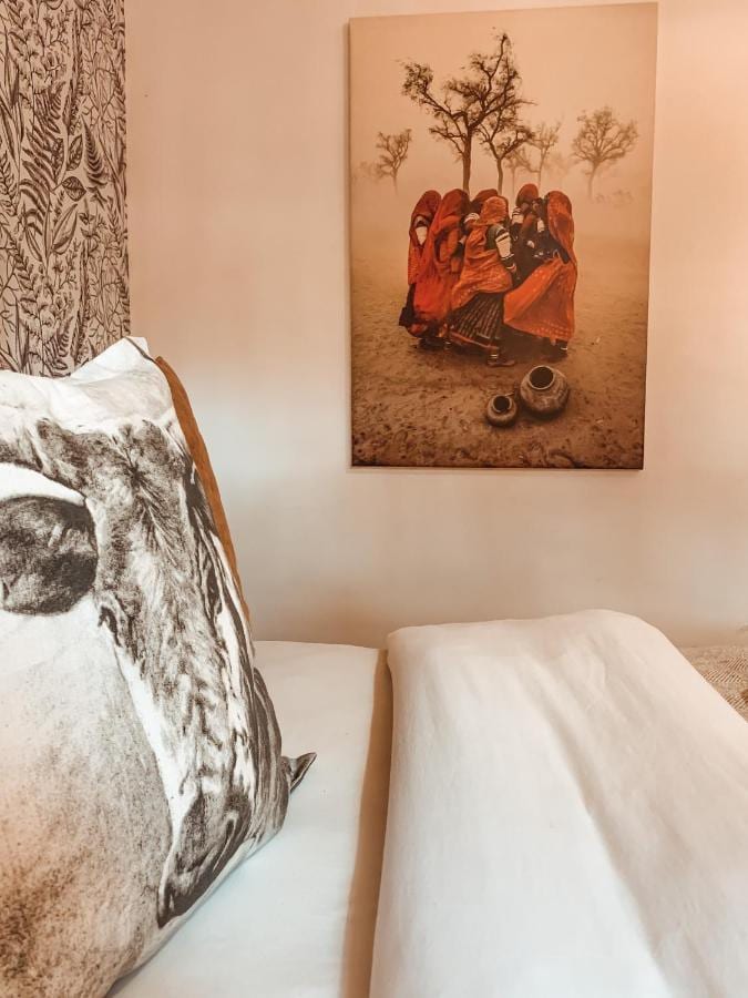 Un coussin à tête de vache sur un lit blanc, sous un tableau de femmes en voiles rouges au pied d'un arbre