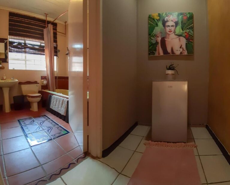 Une salle de bains avec baignoire, lavabo et toilettes, vue depuis un couloir où un petit réfrigérateur est posé sous un portrait de femme