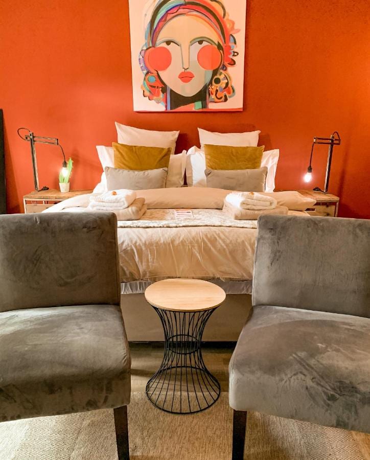 Un grand lit aux coussins moutarde contre un mur orange, sous un portrait coloré, avec deux fauteuils gris en velours et un guéridon