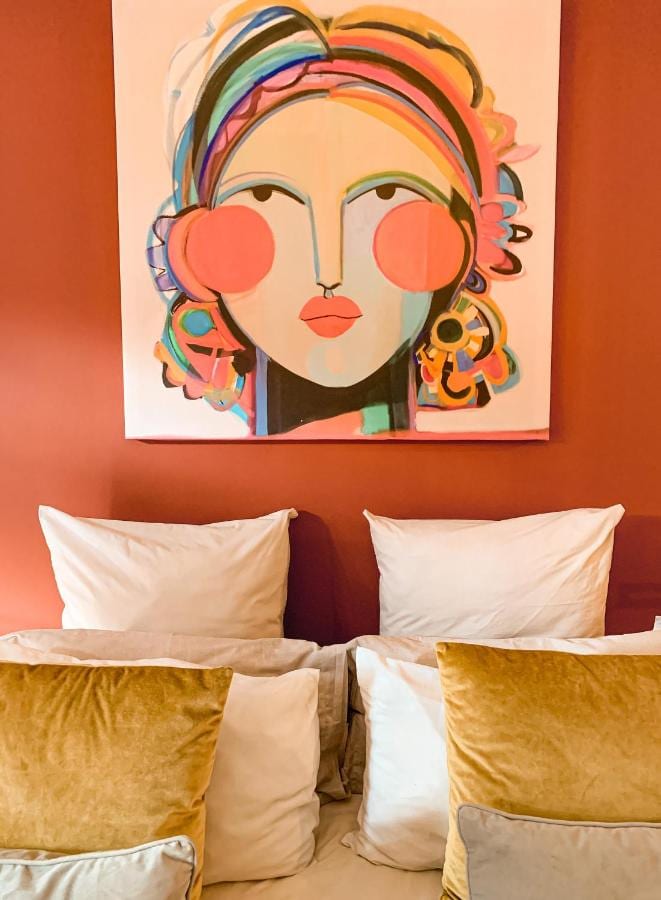 Un portrait de femme aux couleurs vives au-dessus d'un lit aux oreillers blancs et aux coussins moutarde, contre un mur orange