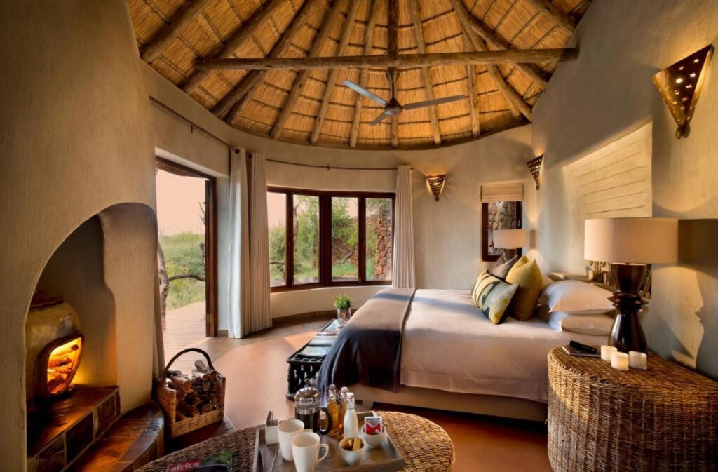 Une suite du Madikwe Safari Lodge sous un toit de chaume, avec un grand lit et une cheminée allumée
