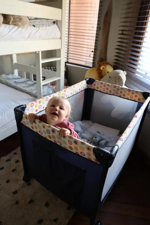 Un bébé souriant dans un lit parapluie, au pied des lits superposés