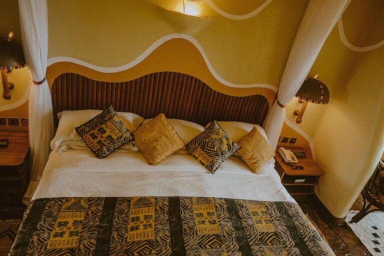 Un grand lit aux coussins à motifs africains, sous une tête de lit en lattes de bois et des murs peints en vagues