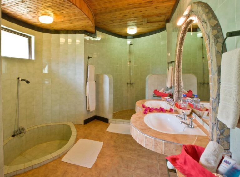 Une salle de bains carrelée de vert, avec un bac de douche rond encastré et deux vasques sous un miroir en arc