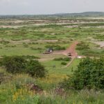 Vue depuis Observation Hill sur une plaine verte tachée de sel, une piste rouge, trois 4x4 garés près d'un petit bâtiment ocre, des collines basses au loin