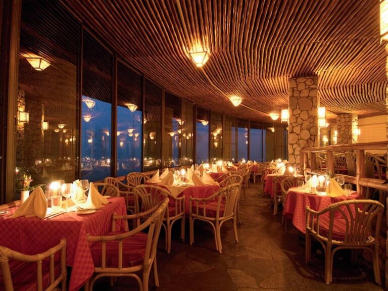 La salle du restaurant éclairée aux bougies, ses nappes rouges et ses fauteuils en rotin sous un plafond en bois