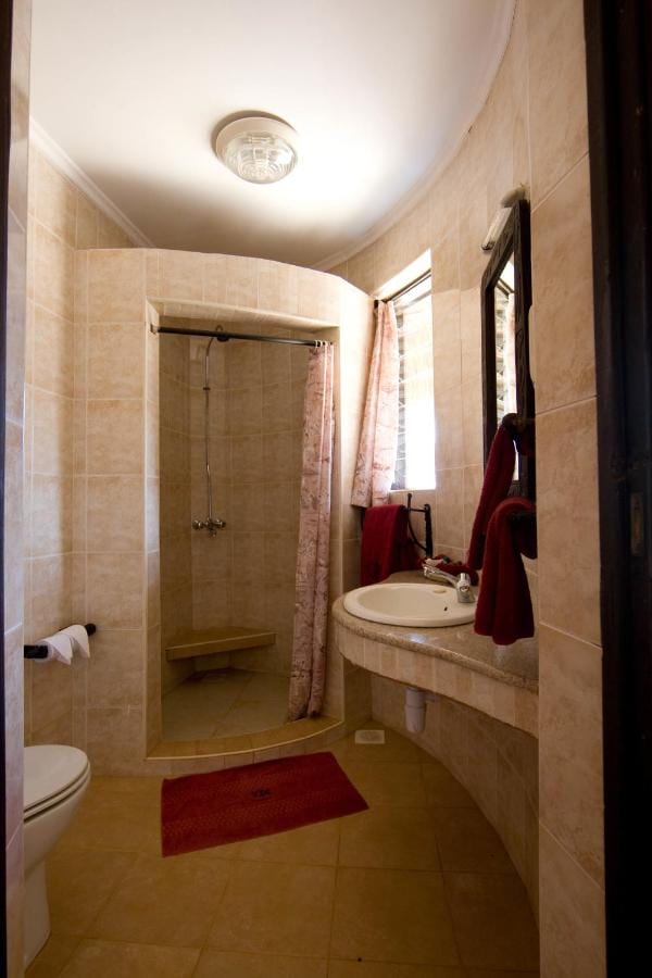Une salle de bains carrelée de beige, avec une douche à rideau, un lavabo sur un plan arrondi et des serviettes rouges