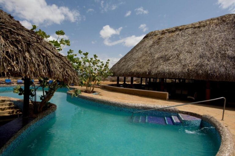 La piscine en mosaïque bleue, entre une paillote et un grand bâtiment au toit de chaume