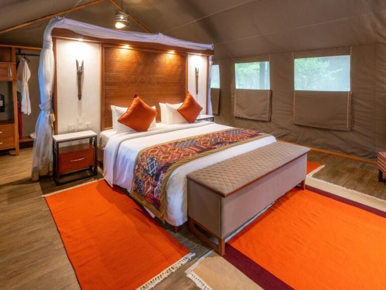 Une tente avec un grand lit sous moustiquaire, des coussins orange et un banc en bout de lit
