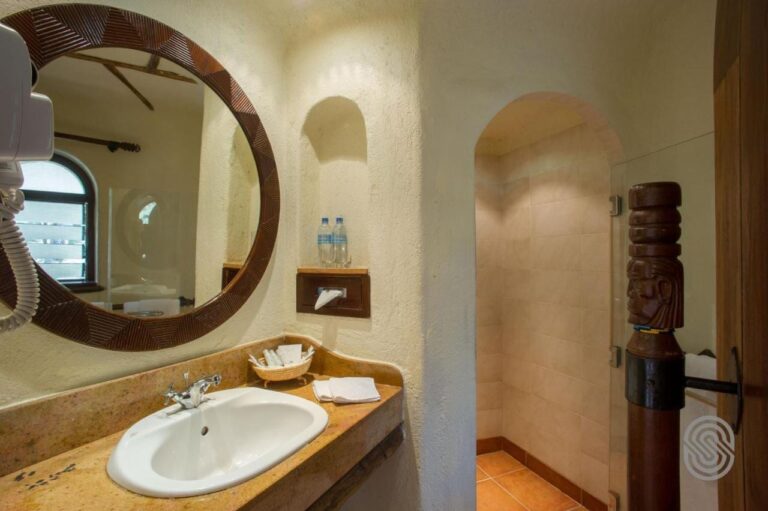 Une salle de bains aux murs crépis, avec un miroir rond cerclé de bois sculpté et une douche sous une arche