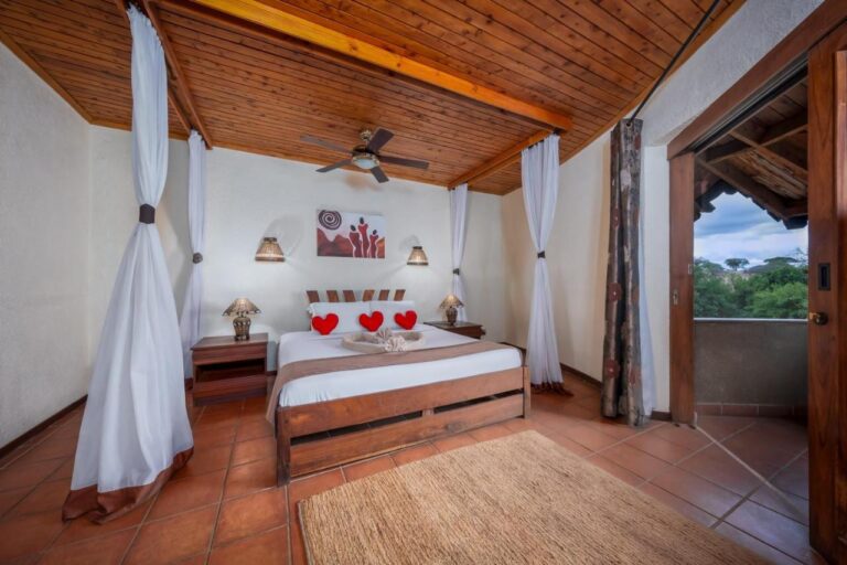 Une chambre au grand lit sous un plafond en bois, avec des voilages et un balcon