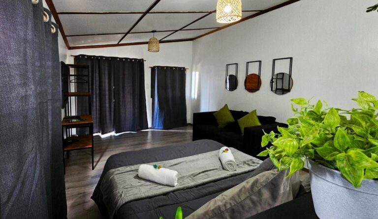 Un grand lit gris avec des serviettes roulées, un canapé noir et des rideaux sombres, dans un studio au plafond quadrillé de bois