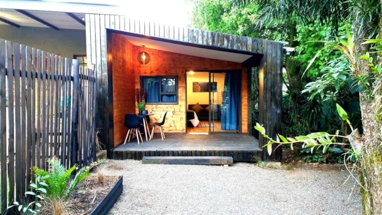 Un chalet de bois avec sa terrasse couverte, une table et deux chaises noires, devant une cour de gravier et des plantes