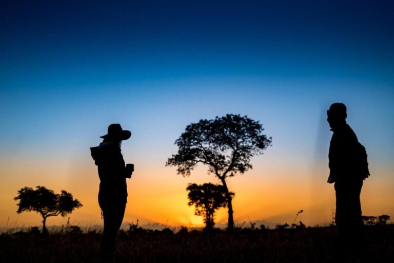 Deux personnes en silhouette dans la brousse, dont l'une tient une tasse, devant des arbres et un horizon orange